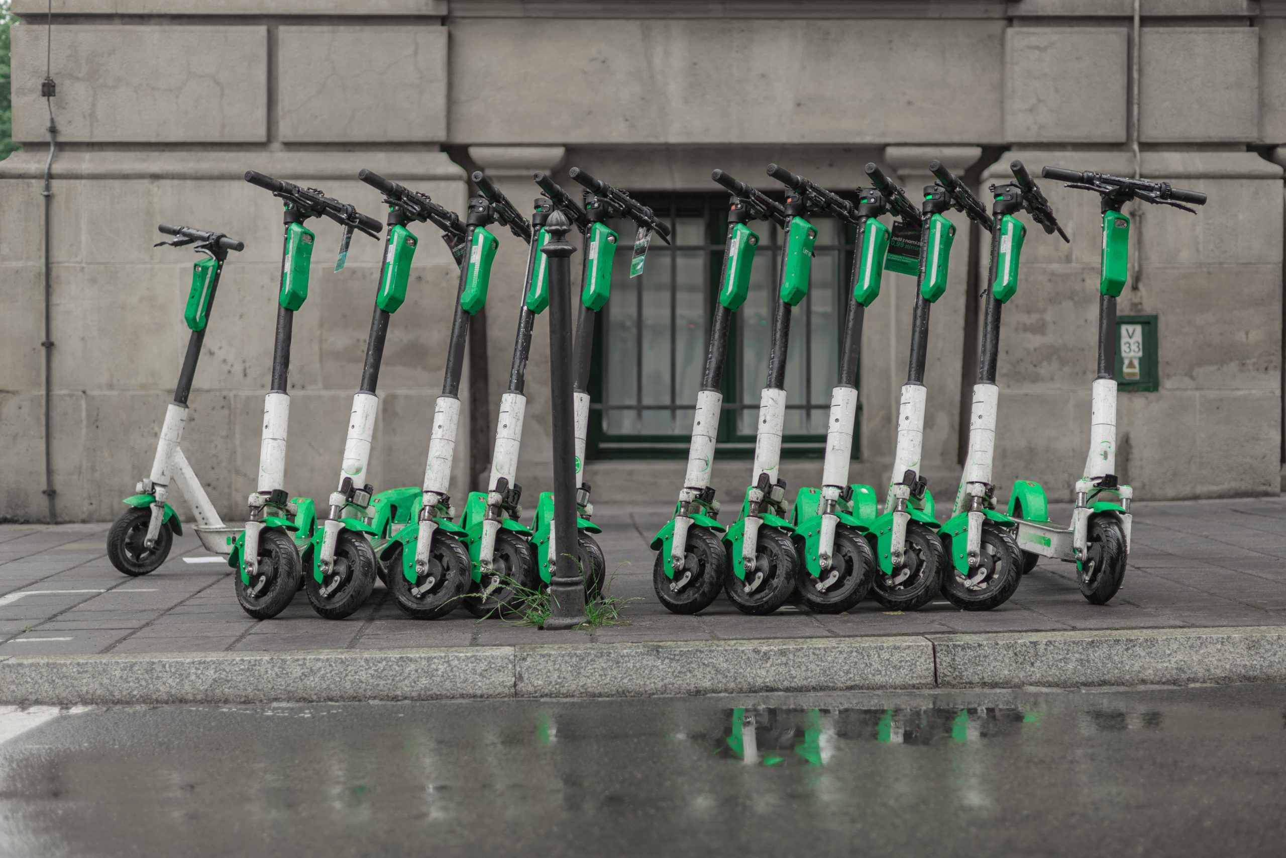 A row of electric scooters in green and white colors is parked along a city sidewalk. The scooters are lined up neatly, with some reflections visible on the wet pavement