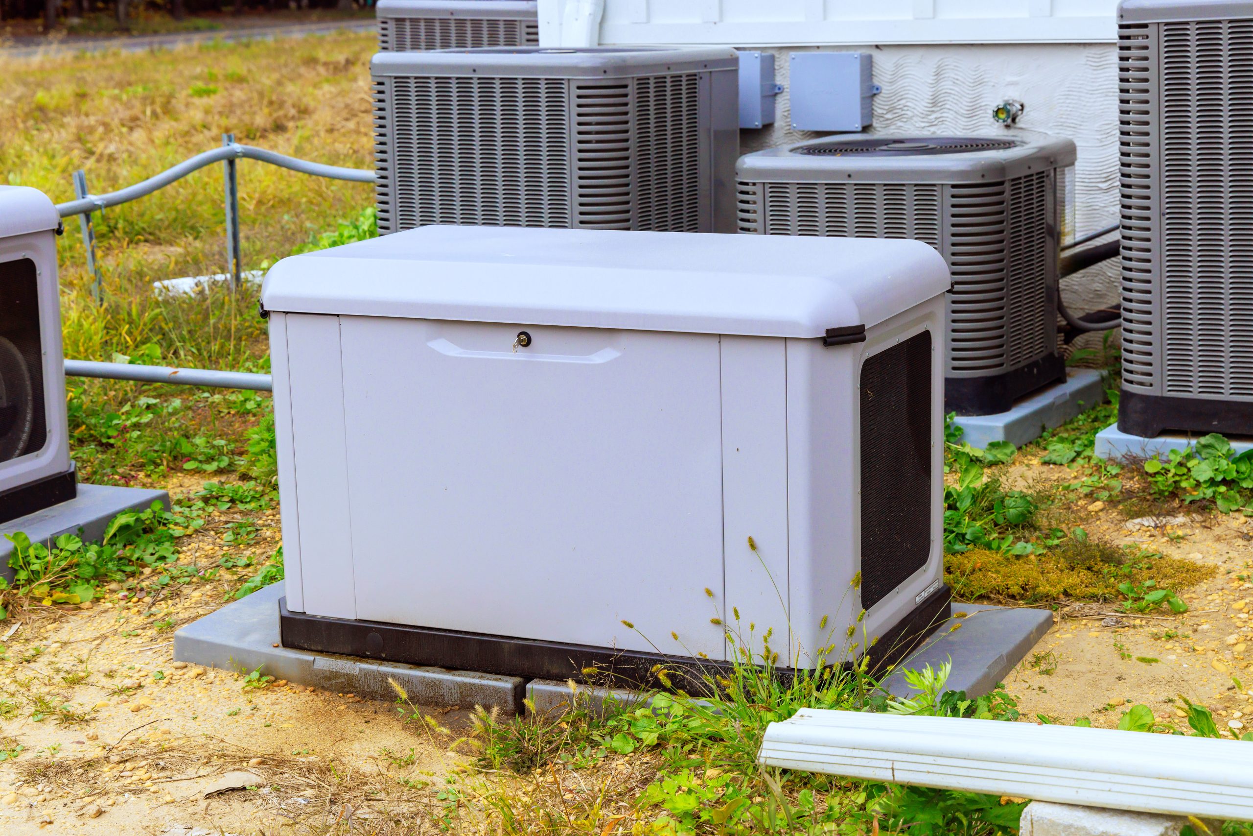 A gray generator sits on a concrete pad in a grassy area, surrounded by several air conditioning units. The generator has a flat top and a small door on the front
