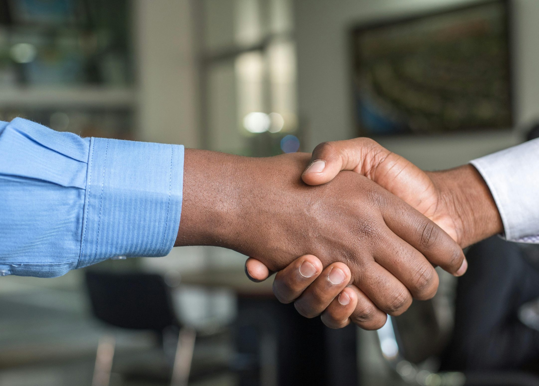 Two hands are engaged in a handshake, one wearing a blue long-sleeve shirt and the other in a white long-sleeve shirt. The background features a blurred office setting