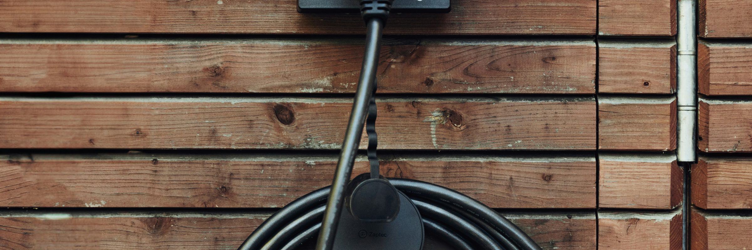 A black electric vehicle charging station mounted on a wooden wall, with a coiled charging cable hanging down. The charging connector is attached to the station