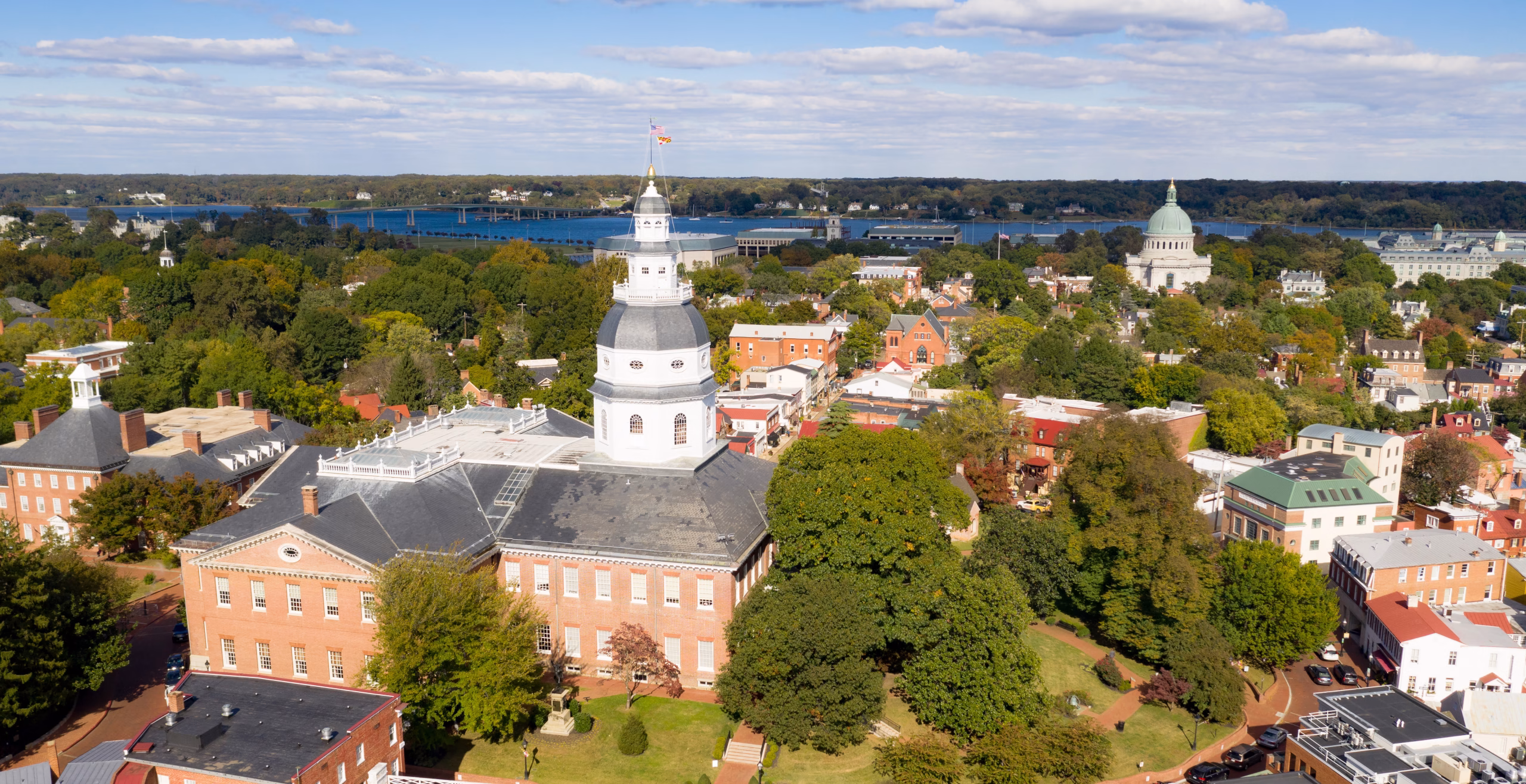 Aerial view of a city featuring a prominent dome-topped building, likely a state capitol, surrounded by trees and residential structures. In the background, a body of water is visible, along with additional buildings and greenery. The scene is set under a partly cloudy sky