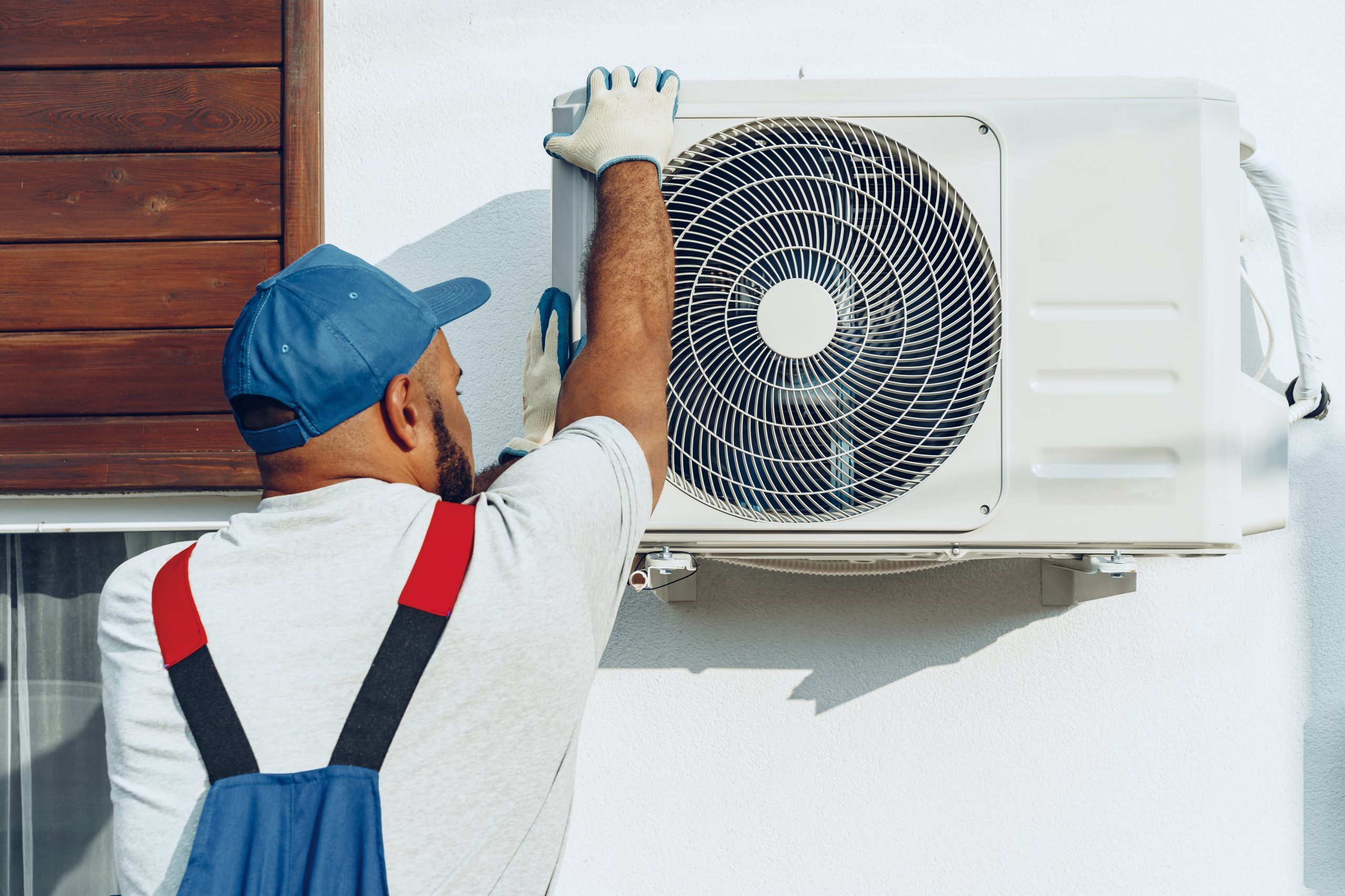 A person wearing a blue cap and gloves is adjusting an outdoor air conditioning unit mounted on a wall. They are dressed in a gray shirt and blue overalls, facing the unit while reaching up to make an adjustment