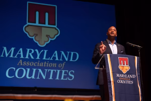 A speaker stands at a podium with a banner displaying the Maryland Association of Counties logo. The speaker gestures with one hand while addressing an audience. The background features a large logo of the organization