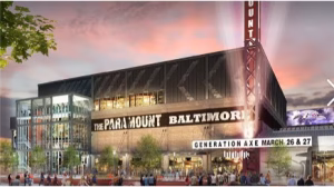 A modern theater building with a large marquee displaying "THE PARAMOUNT BALTIMORE" and event details. The structure features a glass facade and is surrounded by trees and landscaping, set against a colorful sunset sky