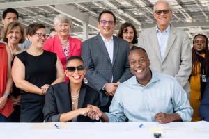 Two individuals are seated at a table shaking hands, smiling at each other. Behind them, a diverse group of people stands.