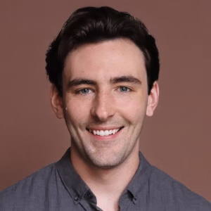 A young man with dark hair and blue eyes smiles at the camera. He has a light complexion with some freckles and is wearing a gray button-up shirt. The background is a solid, warm brown color