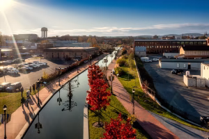 A scenic view of a canal lined with trees displaying autumn foliage. People walk along the pathway beside the water, and buildings are visible in the background. A water tower and distant hills are also part of the landscape, with sunlight illuminating the scene
