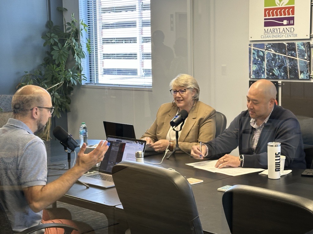 Three people are seated at a conference table in a modern office setting. They appear to be recording a podcast.