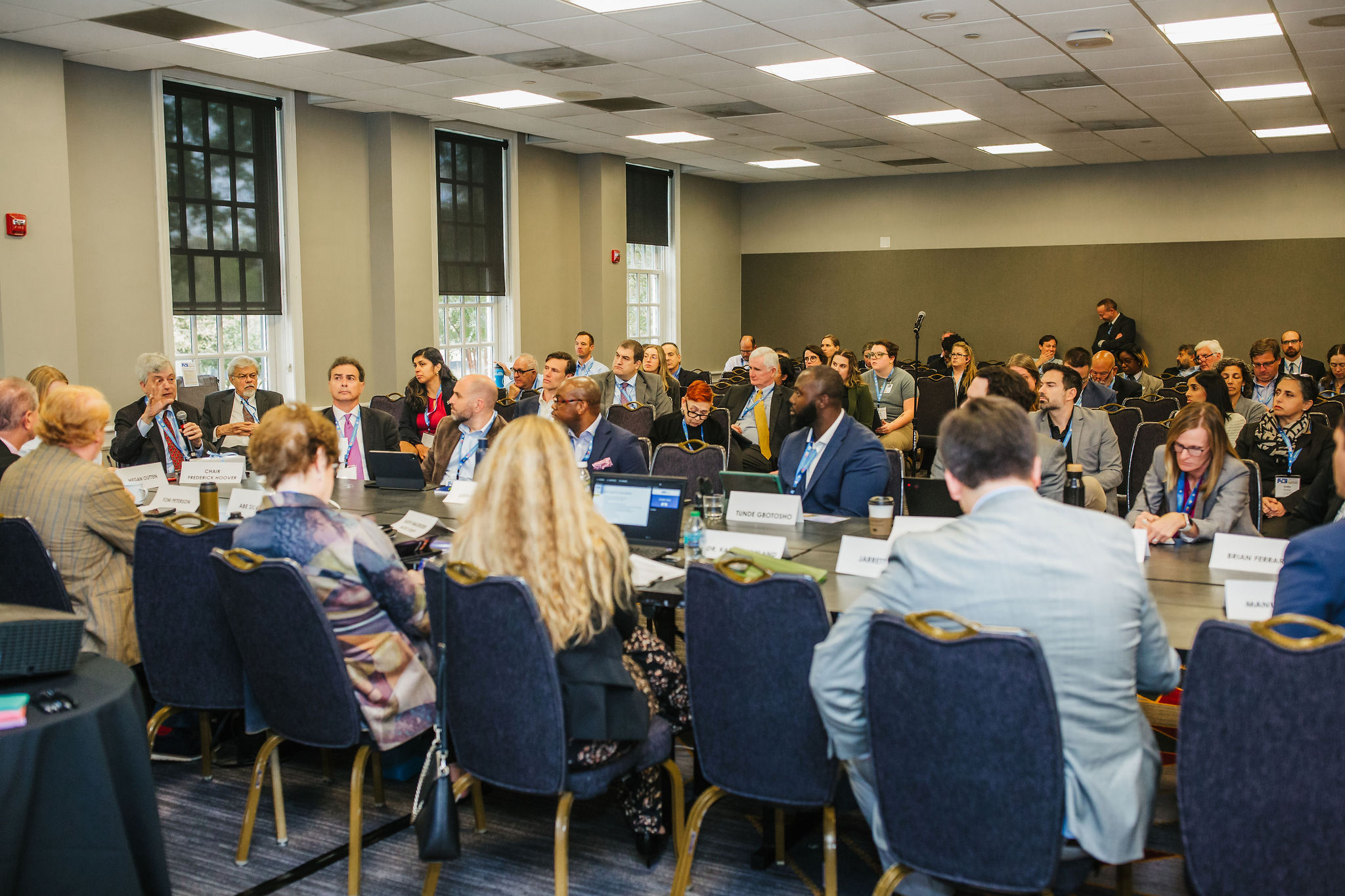 A group of people seated around a large conference table in a meeting room. Attendees are engaged in discussion, with some taking notes and others listening attentively. Nameplates are visible in front of several participants. The room has large windows and a professional setting