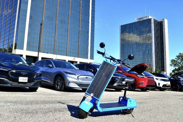 A blue electric scooter with a solar panel on the front is parked in a lot filled with various cars, including several sedans and SUVs. In the background, there are tall glass buildings under a clear sky
