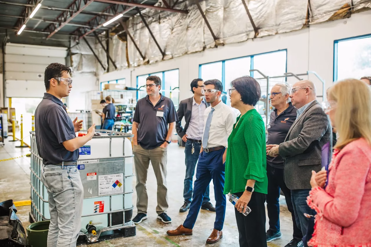 A group of eight people listens attentively to a speaker in a warehouse setting. The speaker, wearing a dark shirt, gestures while explaining something. The audience includes individuals in business attire, with some wearing glasses. The background features industrial equipment and large windows