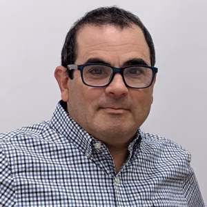 A man with short dark hair and glasses is wearing a checkered button-up shirt. He is looking directly at the camera with a neutral expression against a plain background