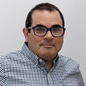A man with short dark hair and glasses is wearing a checkered button-up shirt. He is looking directly at the camera with a neutral expression against a plain background