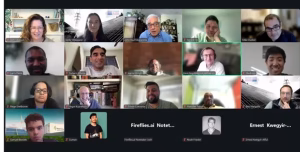 A virtual meeting with multiple participants displayed in a grid format. The screen shows a mix of smiling faces and individuals engaged in conversation, with some people raising their hands. The background includes various home office settings