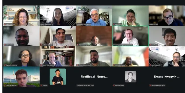A virtual meeting with multiple participants displayed in a grid format. The screen shows a mix of smiling faces and individuals engaged in conversation, with some people raising their hands. The background includes various home office settings
