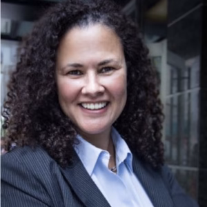 A woman with curly hair smiles confidently while wearing a dark blazer over a light blue shirt. She stands in an urban setting with buildings in the background