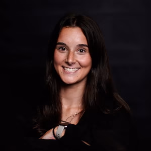 A young woman with long, dark hair smiles while wearing a black top. She has a fitness tracker on her wrist and is posed against a dark background