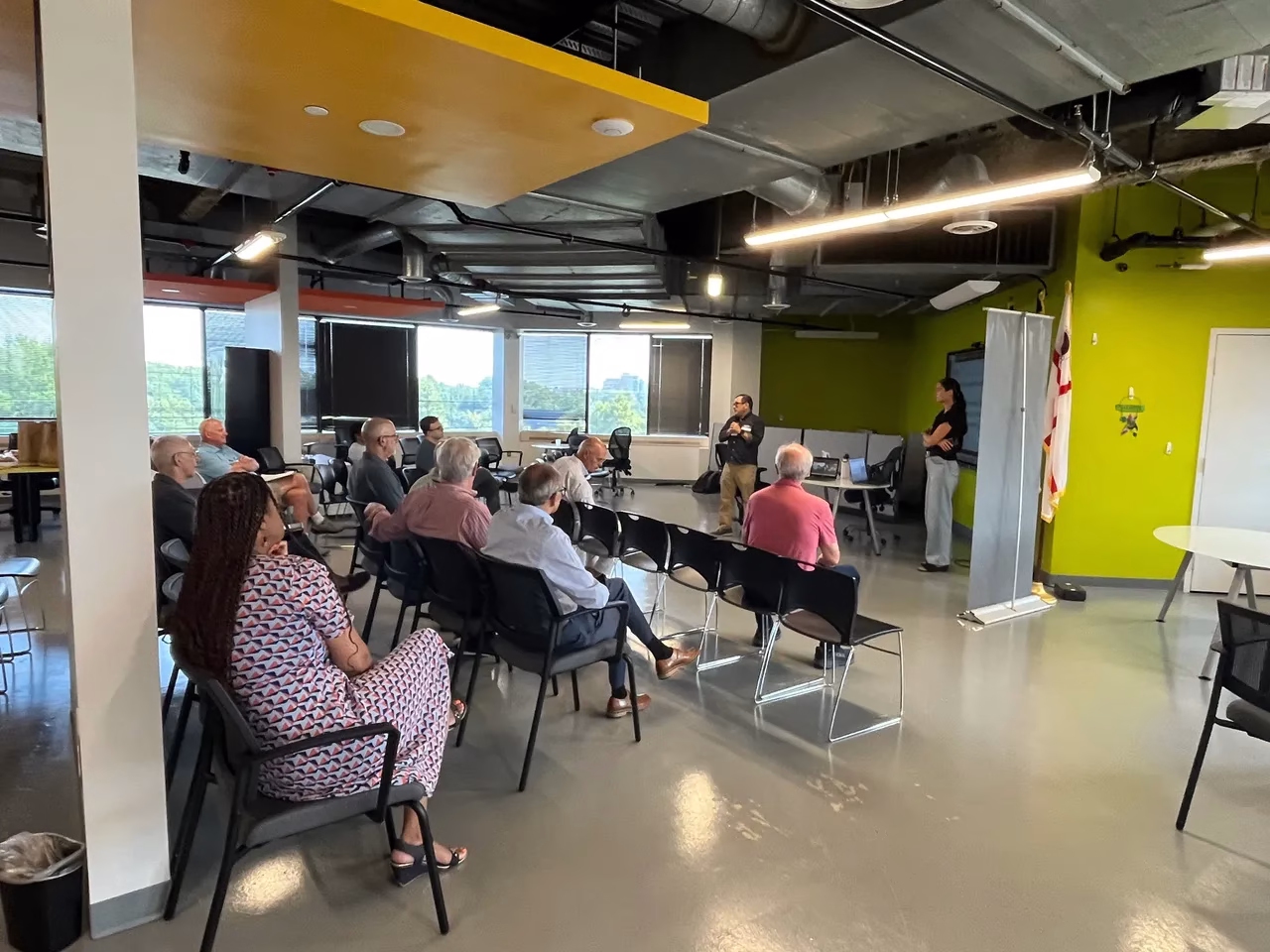 A group of people sits in chairs facing two speakers in a modern indoor space. The audience, consisting of mostly older adults, is engaged in a discussion. The room features large windows with a view of greenery outside, and the walls are painted in bright colors