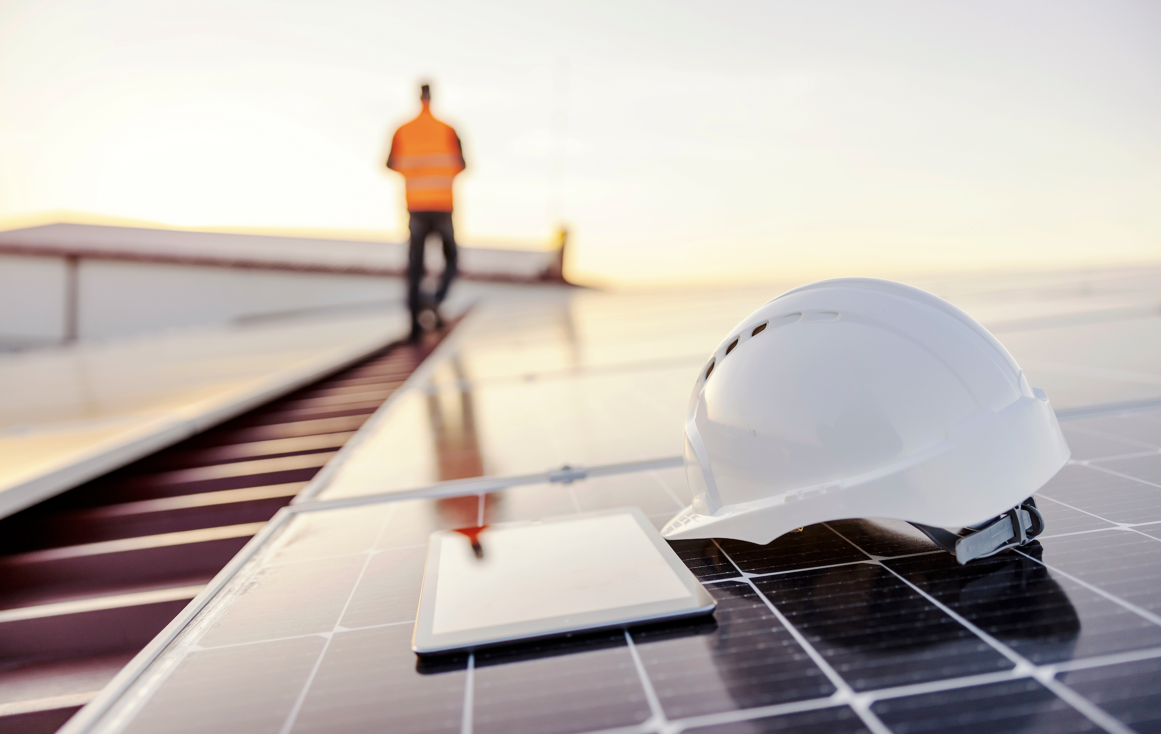 A white hard hat rests on a solar panel, with a tablet beside it. In the background, a person wearing an orange safety vest stands on a rooftop, looking at a device in their hands. The scene is set during sunset, with a clear sky