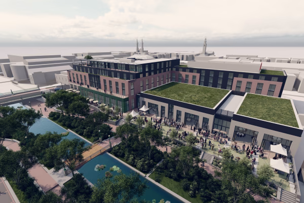 A modern multi-story building with a green roof is situated near a landscaped area featuring trees and a pond. People are gathered outside, likely for an event, with tables and tents set up. The surrounding area includes additional buildings and a clear sky above