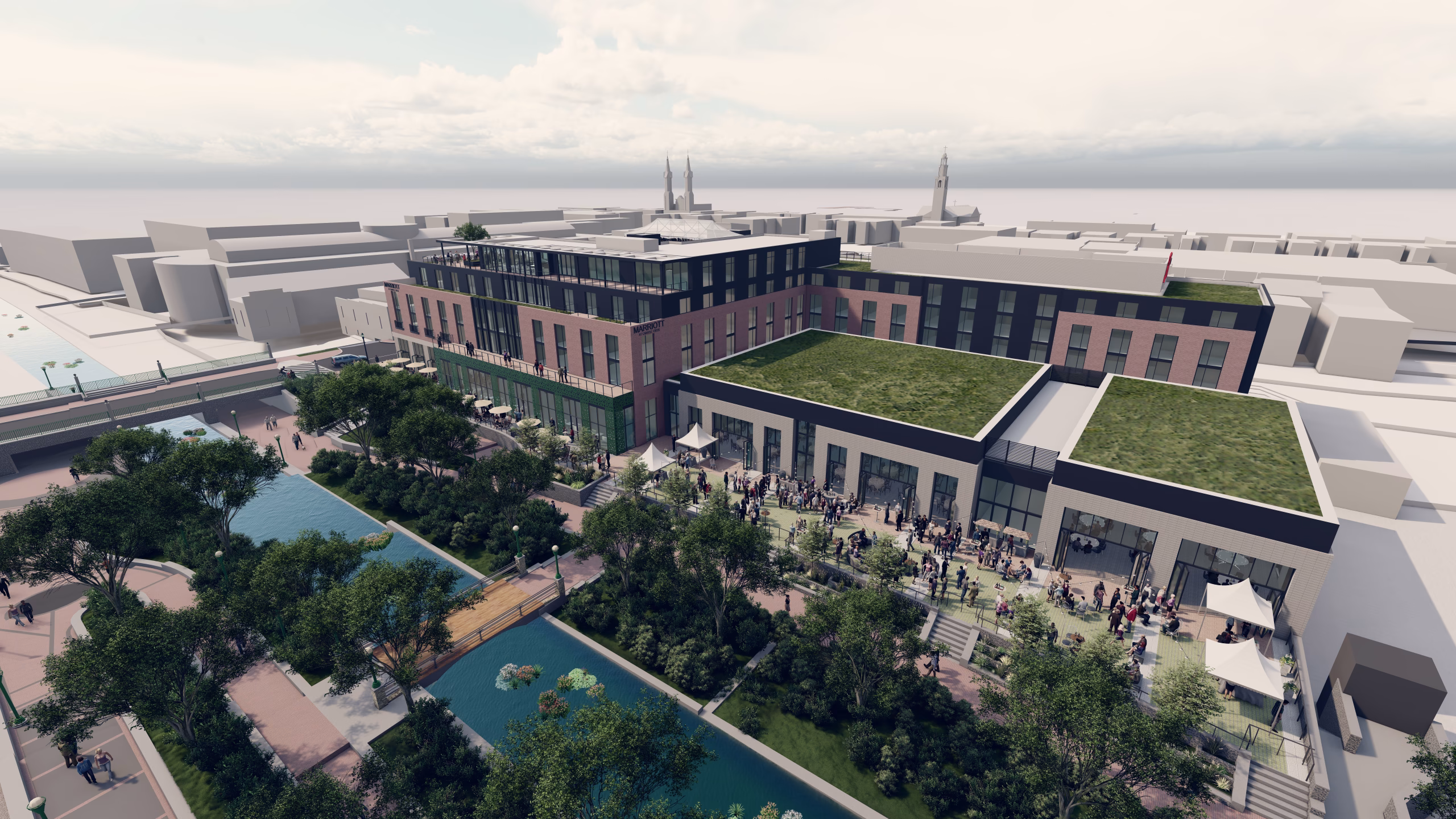 A modern multi-story building with a green roof is situated near a landscaped area featuring trees and a pond. People are gathered outside, likely for an event, with tables and tents set up. The surrounding area includes additional buildings and a clear sky above