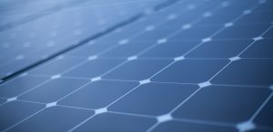 Close-up view of solar panels, showcasing a grid of dark photovoltaic cells with a textured surface and a geometric pattern. The panels are arranged in rows, reflecting light