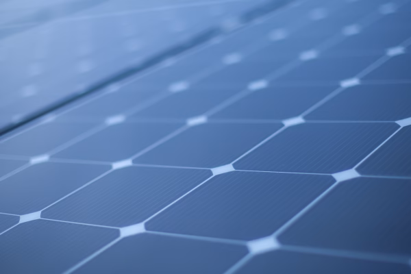 Close-up view of solar panels, showcasing a grid of dark photovoltaic cells with a textured surface and a geometric pattern. The panels are arranged in rows, reflecting light