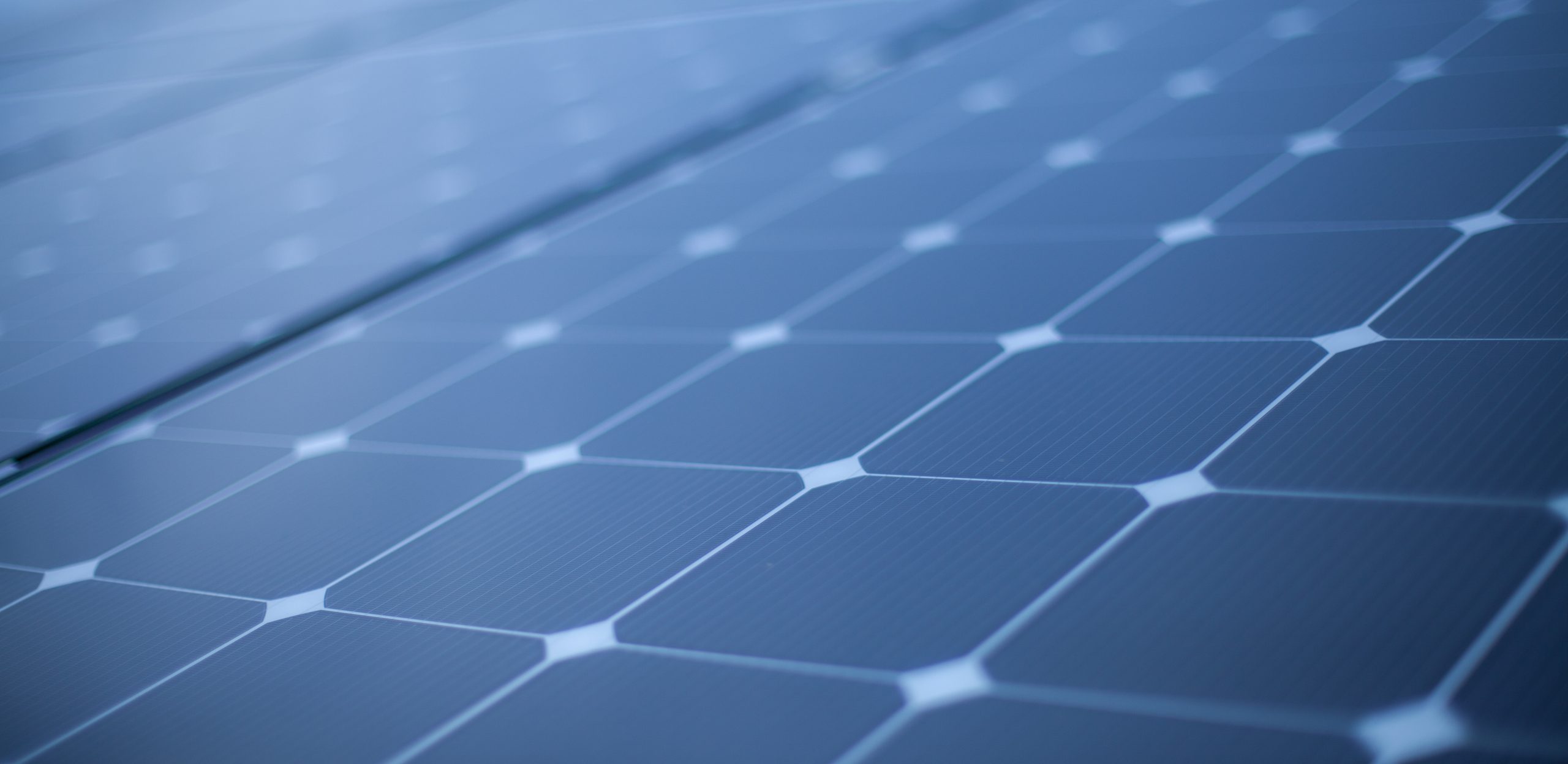 Close-up view of solar panels, showcasing a grid of dark photovoltaic cells with a textured surface and a geometric pattern. The panels are arranged in rows, reflecting light
