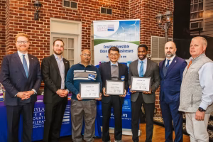 A group of six men stands together, holding certificates. They are dressed in business attire and casual clothing. The background features a brick wall and tables with promotional materials. The men appear to be celebrating an achievement related to clean energy or business innovation
