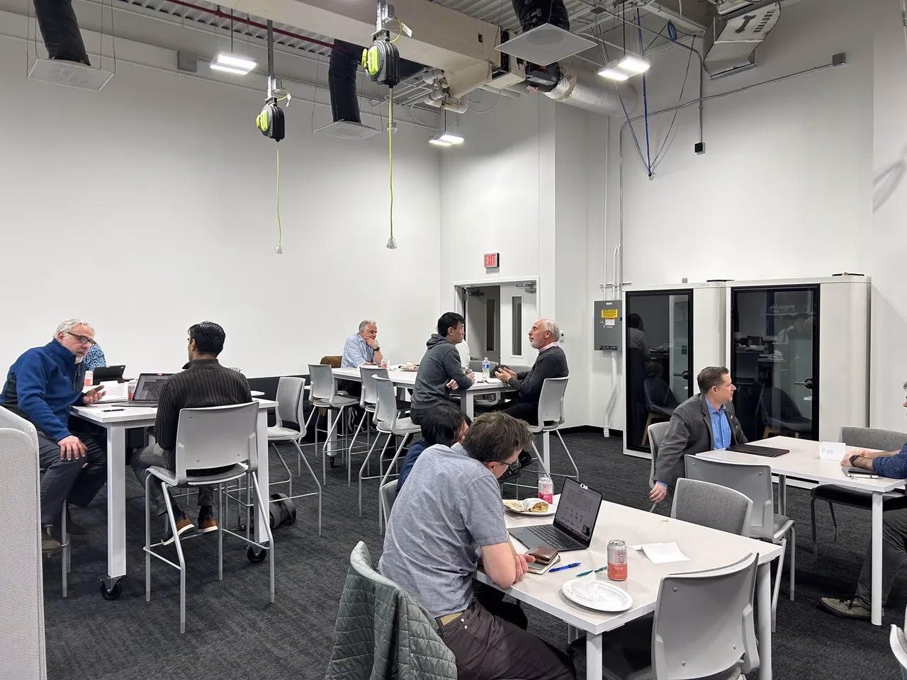 A spacious room with several tables and chairs, where people are engaged in discussions and working on laptops.