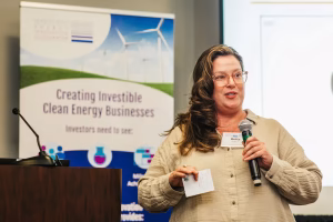 A woman with long, wavy hair speaks into a microphone while holding a notepad. She is wearing a light-colored button-up shirt and stands in front of a banner promoting clean energy businesses. The banner features wind turbines and text about attracting investors