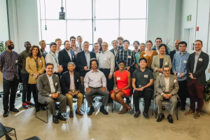 A diverse group of individuals stands and sits in a well-lit room with large windows. They are gathered for a formal event, with some holding awards or recognition plaques. The group includes men and women of various ages and ethnicities, dressed in business attire